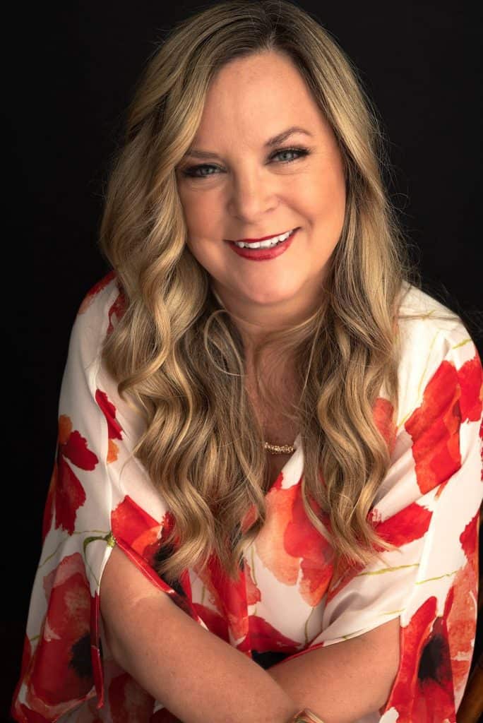 Portrait of a smiling woman with long wavy blonde hair wearing a red and orange floral blouse against a black background.