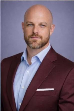 Professional headshot of a bald man in a light blue shirt and burgundy blazer, against a gray background.