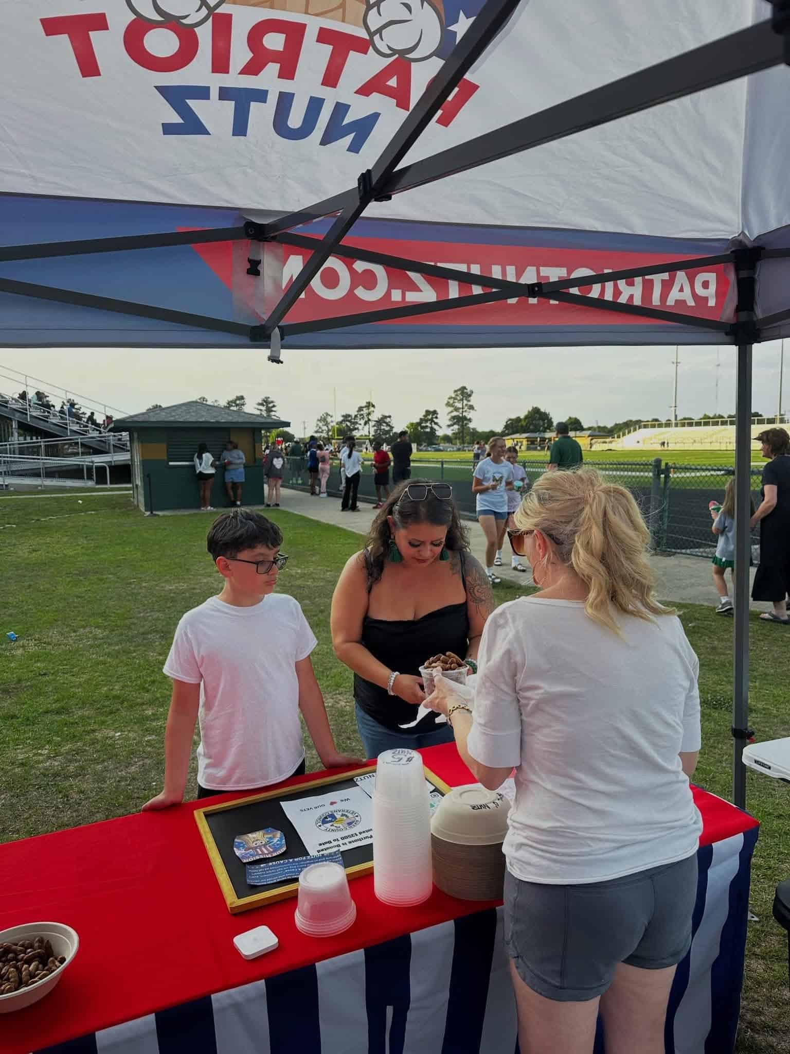 Volunteer table at an outdoor event with a red, white, and blue striped tablecloth; a woman fills cups with nuts while a boy watches nearby.