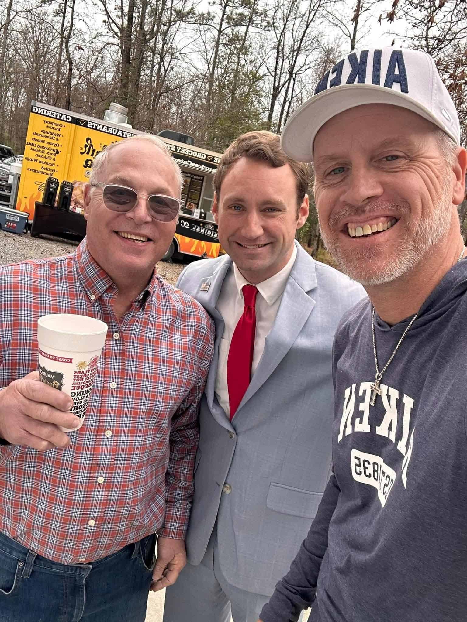 Three men posing for a selfie outdoors, one in a red plaid shirt holding a Starbucks-style cup, another in a light gray suit with a red tie, and a third wearing a cap and dark shirt, with a yellow food trailer in the background.