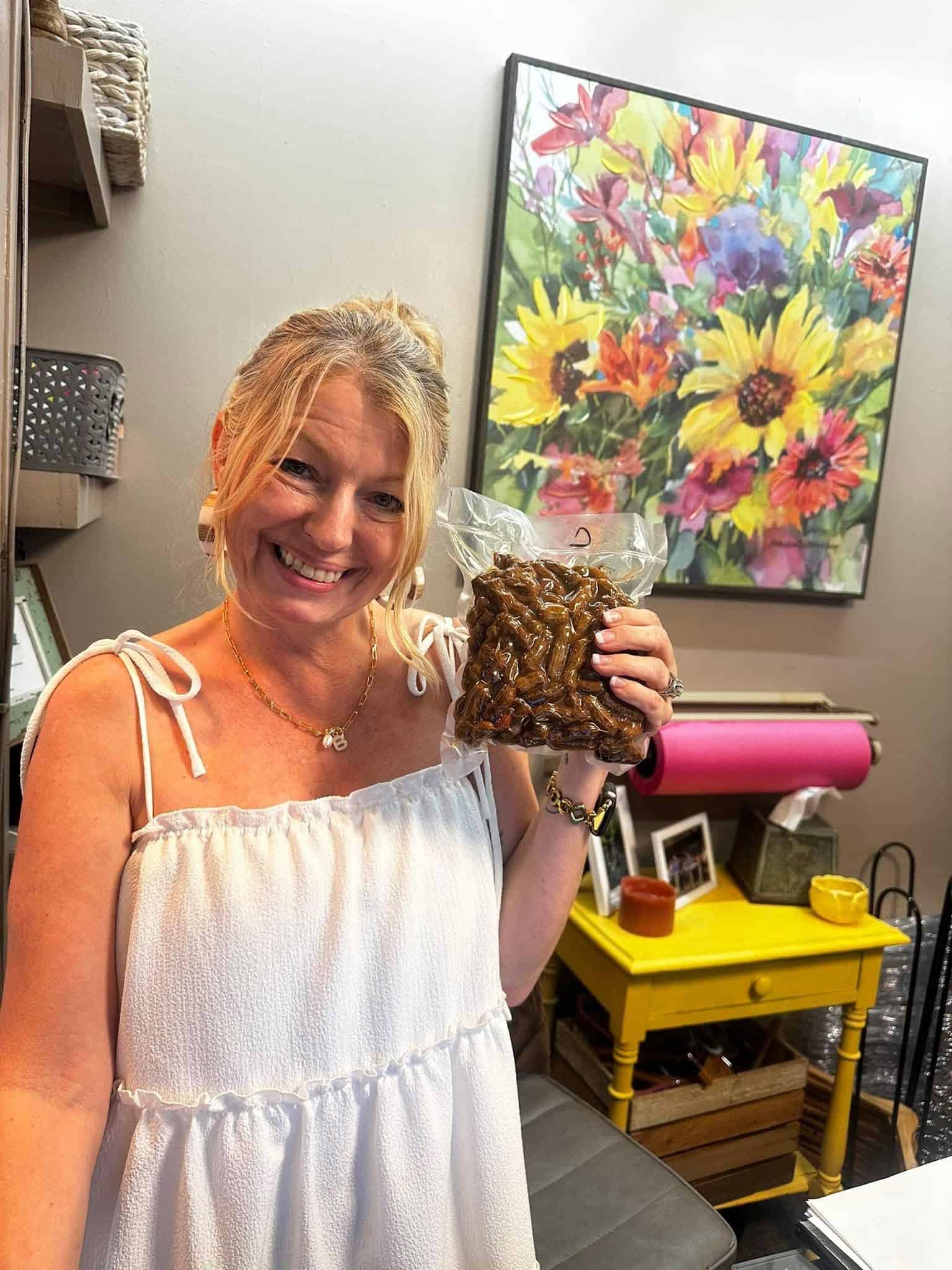 Smiling woman in a white off‑the‑shoulder top holds a large brown pinecone‑like object in a plastic bag, in a room with a bright floral painting in the background.