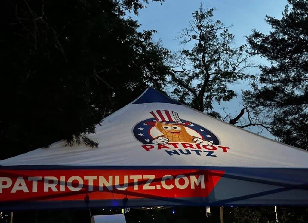 White event tent with the Patriot Nutz logo and the site PatriotNutz.com, set outdoors among trees.