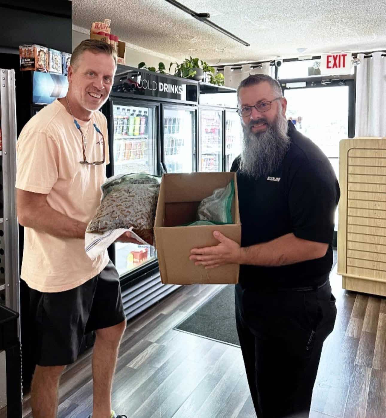 Two men in a store exchange a cardboard box; one holds a garment or towel, the other grins as they hand off the box near a drinks cooler and exit sign behind them.