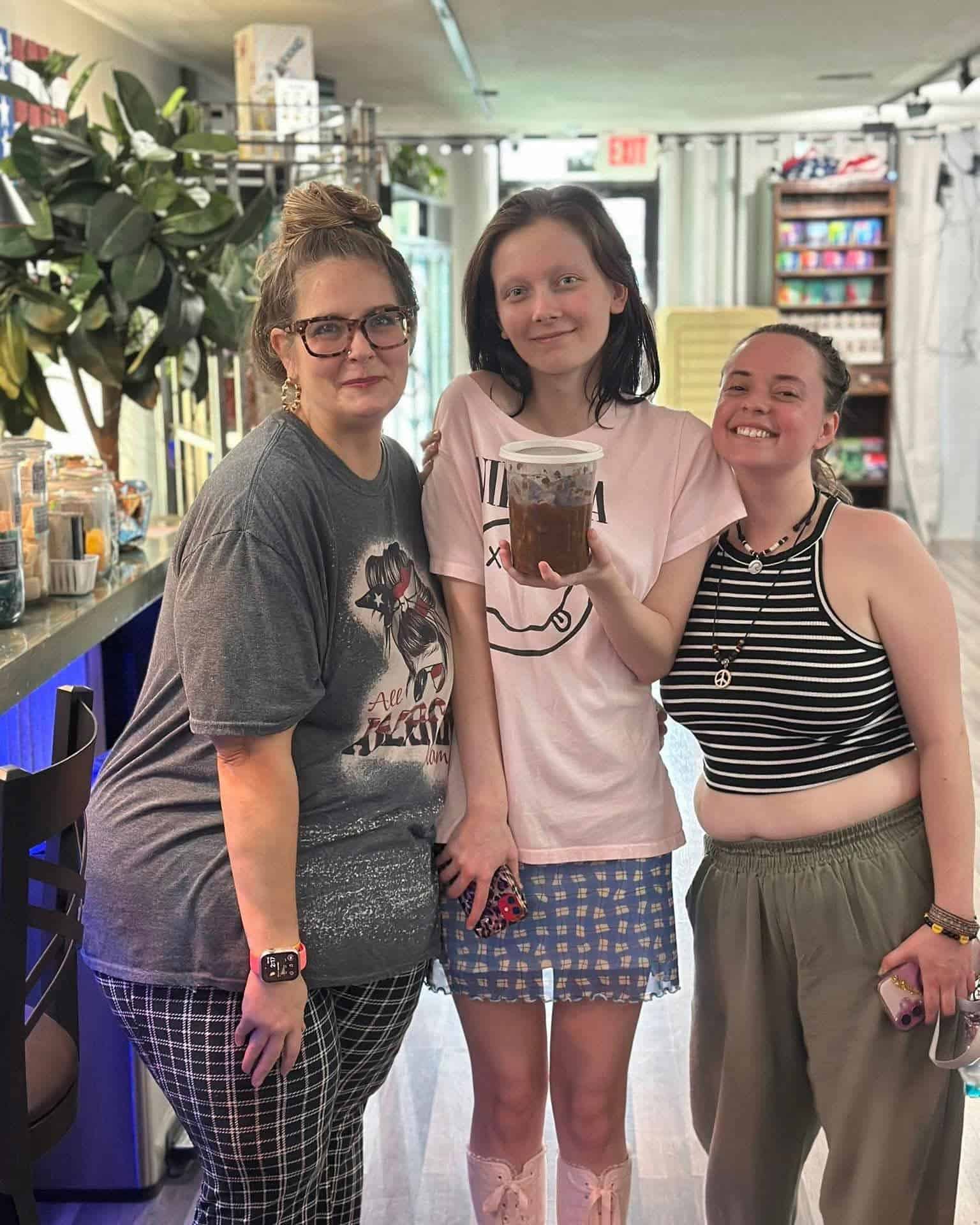 Three friends smiling and posing together in a cafe, with the middle person holding a cup of iced coffee.