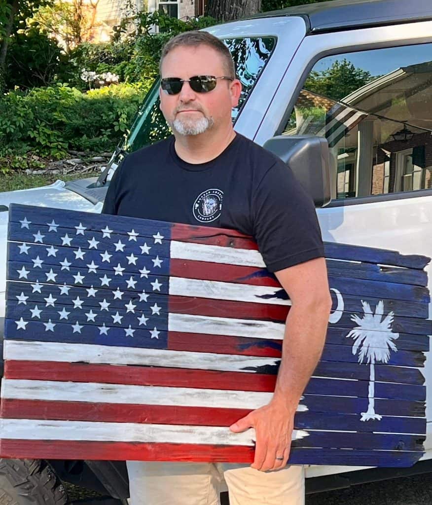Man wearing sunglasses and a black t-shirt stands beside a coffin painted with the American flag design outdoors.