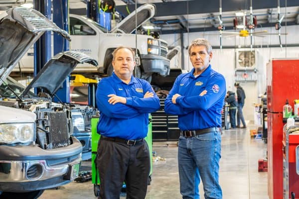 Two auto technicians in blue uniforms stand with arms crossed in a busy repair shop, with cars on lifts and open hoods in the background.