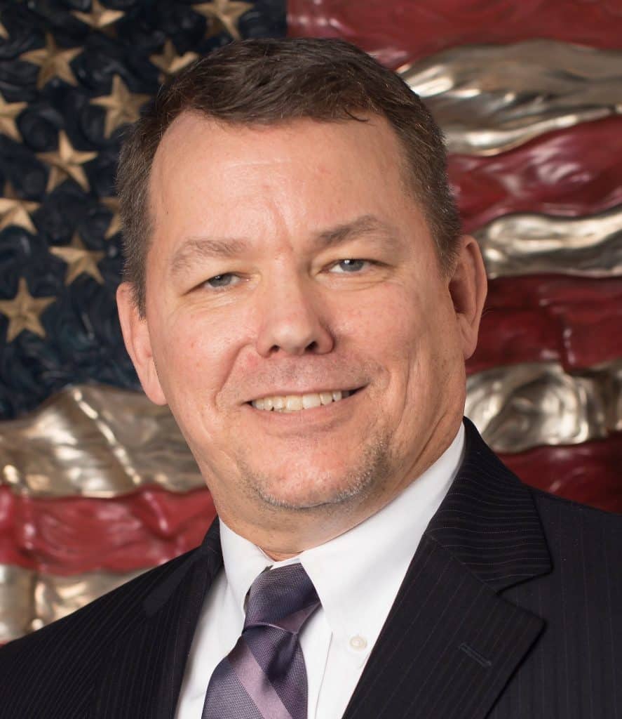 Smiling man in a dark pinstripe suit and lavender tie, with a patriotic flag backdrop behind him.