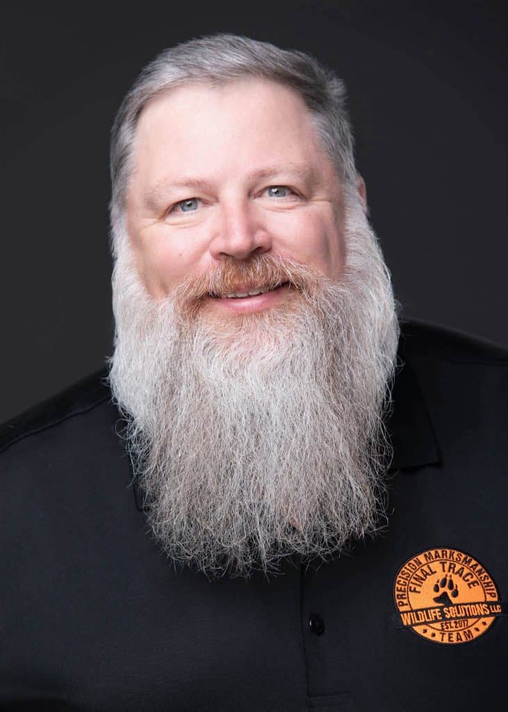 Close-up portrait of a smiling man with a long gray beard and gray hair, wearing a black polo with an orange wildlife‑solutions patch on the chest.