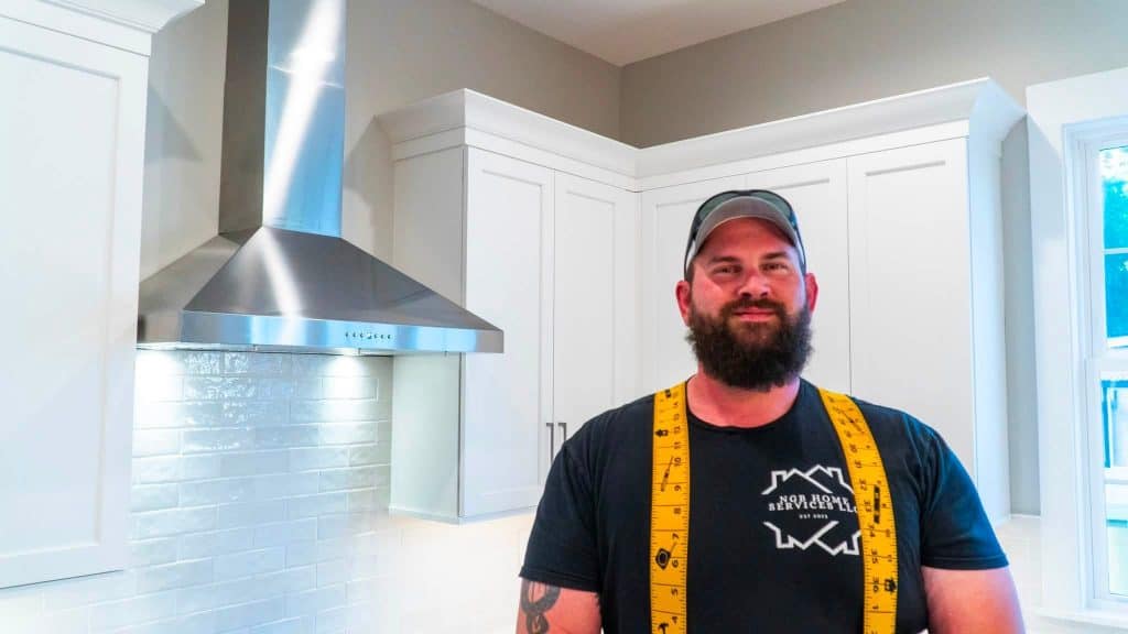 Bearded man in a cap with a yellow measuring tape around his neck stands in a modern white kitchen beside a stainless steel range hood.