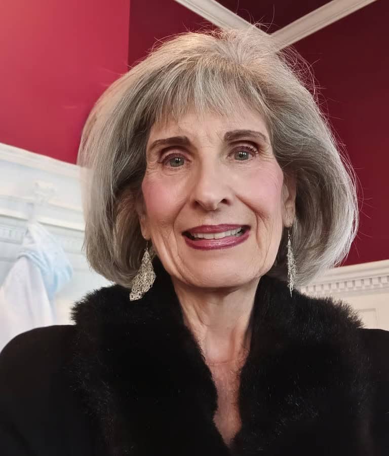 nancy-pic-4-mv0JxzbqzeSW4VVP Smiling elderly woman with silver hair and dangling earrings wearing a black fur-trimmed jacket indoors.