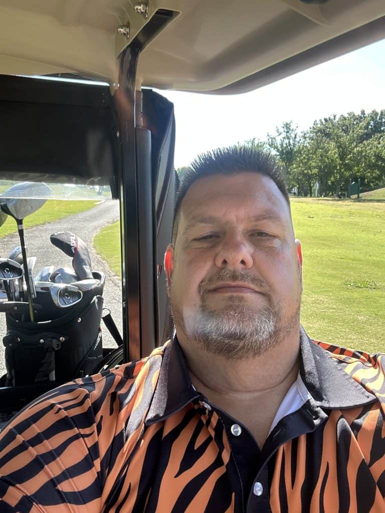 Selfie of a man with short hair and a goatee, wearing an orange/black tiger-stripe shirt, sitting in a golf cart with clubs in view on a sunny golf course.