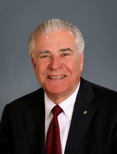 Professional headshot of a smiling older man in a black suit and red tie against a gray backdrop.