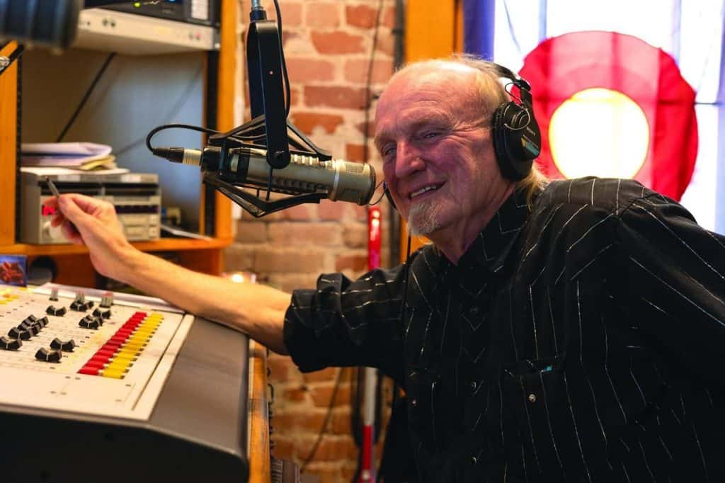 Radio host wearing headphones speaks into a large studio microphone while adjusting a control board in a brick-walled recording studio.