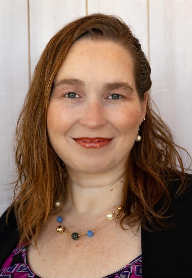 Portrait of a smiling woman with shoulder-length red hair, wearing a black blazer and a colorful beaded necklace.