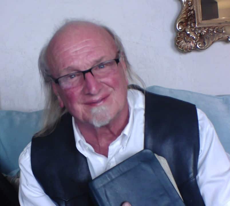 Older man with glasses and a white shirt wearing a black vest sits on a couch, holding a blue book and smiling.